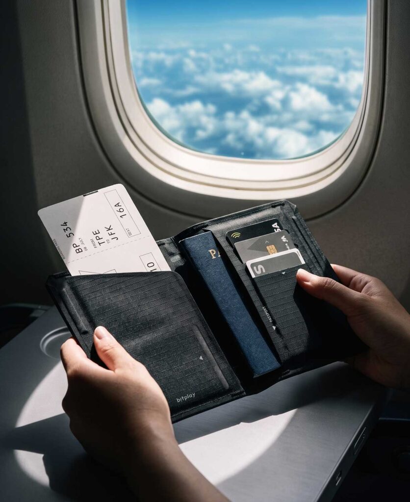 飛行機の窓際の座席で、パスポートと搭乗券を収納したbitplay スリム・バイフォールド パスポートホルダーを開いている様子